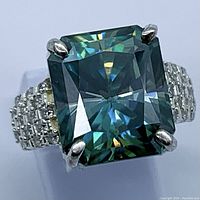 Close-up image of large emerald-cut blue-green moissanite stone set in sterling silver ring with small clear stones along the band.