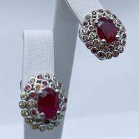 Close-up view of the pair of oval ruby and sapphire earrings displayed on white stands, showing the central oval ruby surrounded by multicolored small round sapphires set in silver with gold plating.