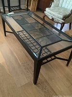 View of the entire coffee table showing the metal frame, glass top and tile insets in center, with surrounding floor and chair.