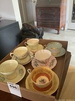 Full lot of six sets of cups and saucers placed in a cardboard box, showing variety in design and color.
