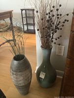 Two ceramic vases on the floor by a wall; one green with round body and narrow neck, the other gray with geometric pattern; both hold dried foliage.