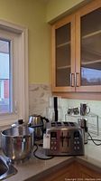 Wide angle photo of all kitchen appliances and cookware on a kitchen countertop near a window with natural light.