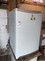 White Whirlpool mini fridge with magnetic stickers on the door, placed on a wooden platform in a basement setting with concrete and cinder block walls.