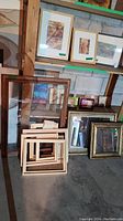 Wide view showing multiple wood frames, some with glass, stacked and leaning against shelving in basement setting.