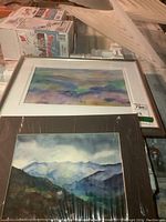 Two framed watercolour paintings on a table. One painting is an abstract or impressionistic landscape titled Bermuda Triangle by MS Rankin with pastel on silk in a silver frame with white matte; the other is a mountain landscape by Billings in watercolour and ink with dark blue and green tones, also framed.