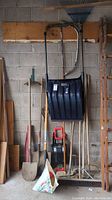 Full view of the tool collection including shovels, post driver, snow shovel and brooms against a cinder block wall.