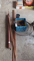 Full view of copper pipes in various lengths with a blue crate containing smaller fittings and a thick red-coated pipe section on concrete floor.