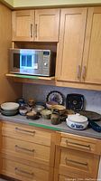 Wide angle photo of kitchen counter displaying assortment of pottery items including bowls, plates and casserole dish beneath wooden cabinets and a microwave.