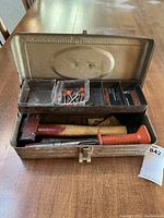 Metal toolbox with hinged lid containing tools and fasteners. Shows the power actuated tool, hammer, and boxes of fasteners inside.