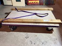 Side angle view of the rolling cart showing wooden planks with reinforced edges, purple strap, and swivel wheels on basement floor.