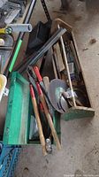 Wood toolbox with green metal toolbox, branch cutters, saw and grinder blades, metal frame pieces and assorted hardware inside.