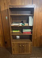 Front view of the veneer bookshelf showing three shelves containing books and one white angled bookend, plus a two-door cabinet at the bottom.