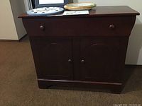 Front view of painted wooden cabinet showing two doors and a single drawer with wooden knobs, dark brown color with scratches and wear visible.