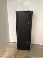 Full front view of a tall black metal cabinet standing in a corner on a concrete floor and next to a white wall.