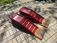 Top and front angled view of the two red metal car ramps mounted on wooden boards placed on a paved ground surface outdoors.