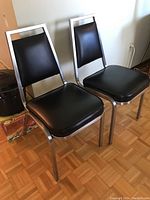 Two black vinyl padded metal framed chairs shown from a frontal angle on parquet floor with some wear, positioned next to wall and other household items.