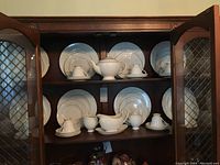 Full view of two shelves in cabinet showing arrangement of Wedgwood china including 8 place settings, teapot, gravy boat, creamer, sugar bowl, cups and plates.