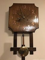 Front view of wooden wall clock showing square face with gold-tone numbers, clock hands, pendulum and key attached.