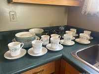 Wide view of dishes including plates stacked, white cups on saucers with floral pattern, and clear glass cups arranged on kitchen counter.