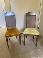 Pair of wooden dining chairs with cane backs and two different colored velvet seat upholsteries, shown against a beige wall and carpet.