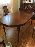 Oval wooden dining table with glossy finish, showing full length with legs, chairs, and parquet floor background.