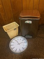 Photo of the analog wall clock, larger metallic brown garbage pail with foot pedal, and smaller beige plastic garbage pail together on carpet