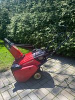 Side angle photo showing whole snowblower with red body, black handles, and wheels on stone paved ground outdoors.