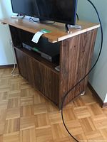 Full view of wooden media stand with flat top, open middle shelf containing a media device placeholder, and lower cabinet doors. Positioned on parquet floor against a wall, with some cables visible.