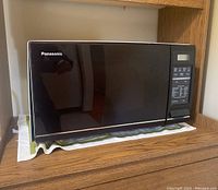 Front view of Panasonic black microwave on wooden shelf with cloth underneath.
