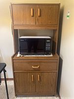 Front view of the tall wood veneer microwave stand with upper cabinet, middle shelf holding a microwave, lower drawer, and cabinet doors.