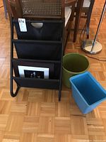 Photo of the magazine rack with black fabric compartments showing the framed photo print inside. Two garbage pails are placed beside the rack, one green and round, the other blue and rectangular.