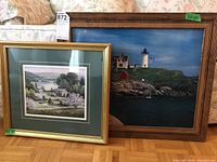 Two framed prints leaning against wall on floor; left is a rural scene, right is lighthouse scene.