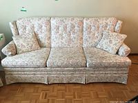 Full view of floral patterned sofa with three cushions and two matching floral throw pillows on a parquet floor near a beige wall.
