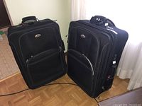 Photo showing two black upright suitcases side by side on wood floor near window with white curtains.