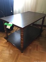 Small wooden side table with turned legs and lower shelf, shown beside a black bag and curtain background.