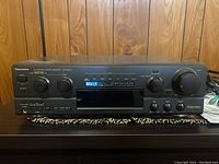 Front view of the Technics SA-AX530 stereo receiver showing control knobs, display, and various buttons, with wood panel background.