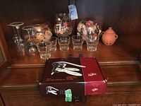 Photo showing two clear glass vases with potpourri, three stemmed wine glasses upside down, four small clear drinking glasses, boxed True Bell wine opener, and orange clay garlic holder on wooden shelf.