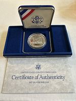 Silver dollar coin in protective capsule displayed inside blue velvet-lined presentation box with patriotic ribbon motif. Certificate of Authenticity laid in front of box.