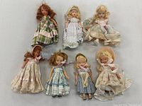 Seven small vintage dolls arranged in two rows on a neutral surface, showcasing a variety of dresses and hair colors.