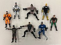 Nine small Batman and one Robin plastic action figures in various colorful Bat suits and poses laid out on white background.