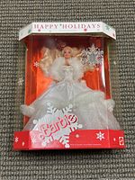 Front view of Barbie doll in original packaging wearing white holiday gown, fur trim, feather wings, and snowflake accessory.