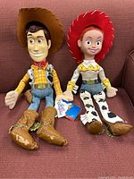 Full view of Woody and Jessie dolls sitting side by side on a red cushion, showing outfits and facial features.