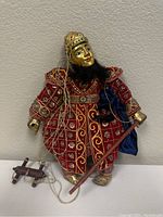 Full view of the Thai style string puppet with the red and gold costume, gold face, black beard, and wooden control bar.