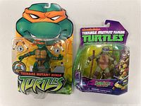 Photo showing Michelangelo and Donatello Teenage Mutant Ninja Turtles action figures in original blister packaging. Michelangelo's pack features an orange mask figure and accessories. Donatello's pack has a purple mask figure with a staff and vibrant packaging.