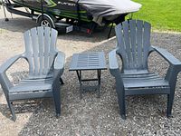 Wide shot showing 2 gray Adirondack chairs and 1 matching folding table outdoors on gravel.