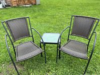 Two rattan and iron chairs with woven seats and backs, seated on grass with a small square side table with a glass top between them.