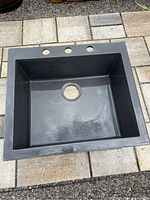 Black granite kitchen sink placed outside on paving stones, visible three faucet holes and drain hole. The sink dimensions and material visible.
