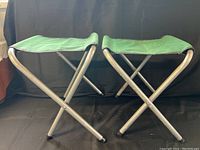 Side view of two green camping stools showing their silver metal tubular frames forming an X shape and fabric seats.