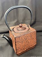 Front and side angled view of square cast iron teapot showing textured reddish-orange surface, black smoothly curved spout and handle, and lid with knob.