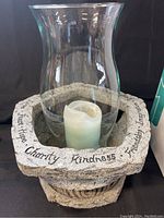 Front view of the stone outdoor candle showing engraved words on top edge, clear hurricane glass, and white pillar candle inside.